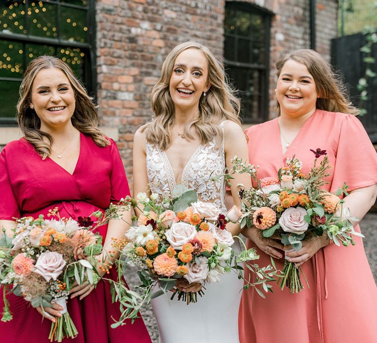 Bride stands with her bridesmaids in mismatched Rewritten Bridesmaid dresses holding orange and white floral bouquets 