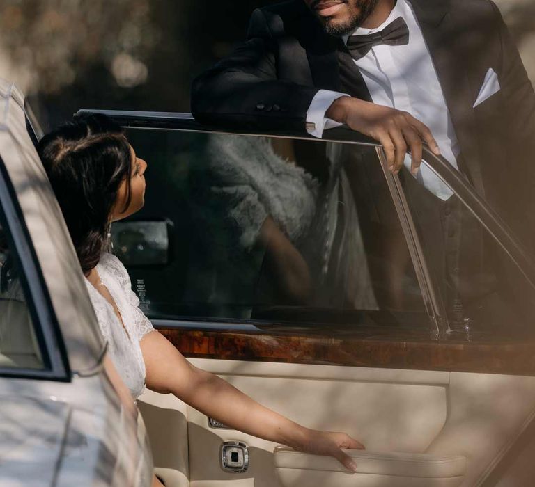 Groom in classic black three piece tuxedo helping bride out of white classic wedding car 