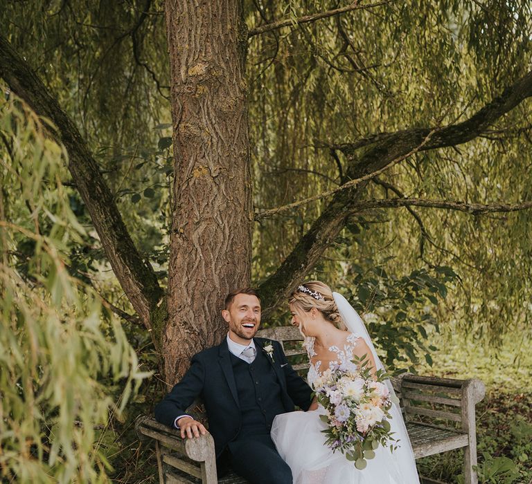 The bride in an illusion lace and tulle wedding dress with sparkly silver shoes sits on a wooden bench laughing with the groom in a navy suit 