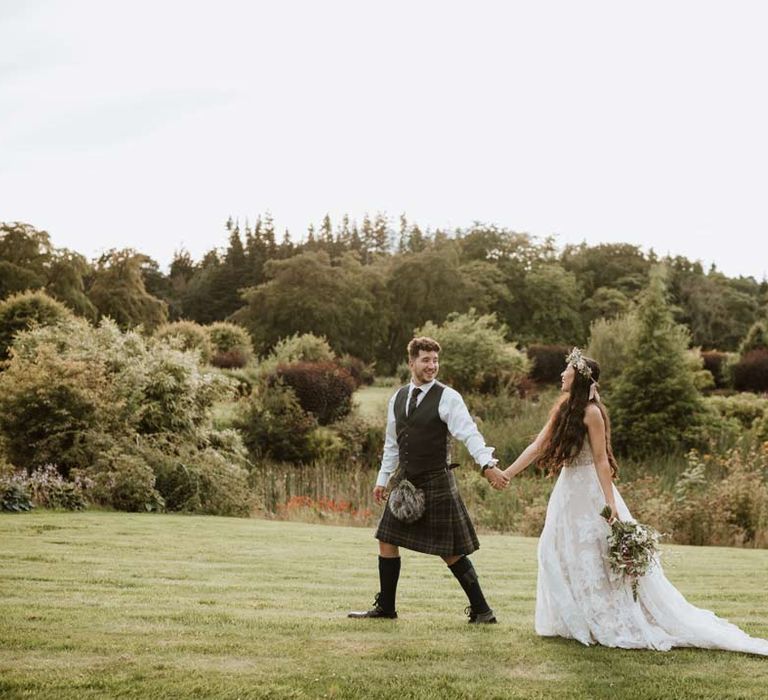 Beautiful wedding couples portraits - groom in dark grey waistcoat, dark tartan tie with white flower boutonniere, dark grey and blue tartan kilt and wedding sporran leading bride wearing celestial 3d appliqué sheer overlay wedding dress and bridal flower crown holding large white rose, peony, dried flower and foliage bridal bouquet 