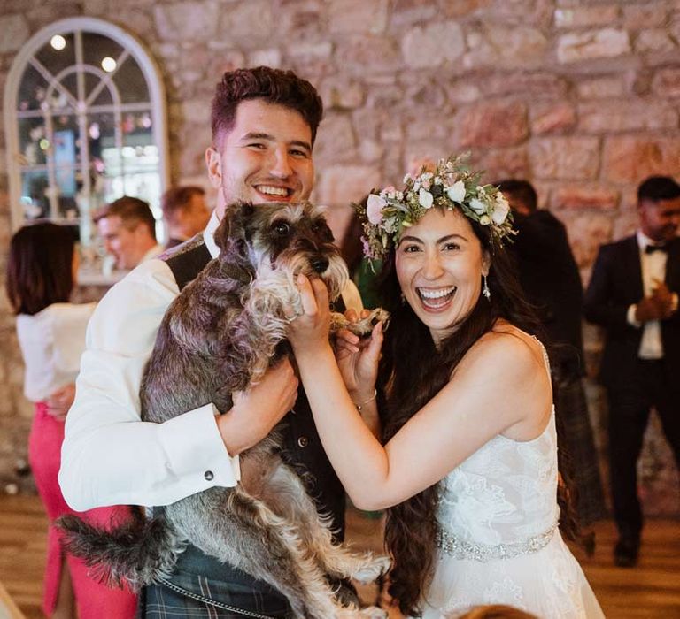 Bride in 3d applique floral wedding dress and groom in dark grey waistcoat and kilt holding up miniature schnauzer puppy at The Barn at Harburn wedding 