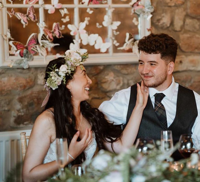 Bride in white garden rose bridal flower crown sitting with groom in dark grey waistcoat and tartan tie at The Barn at Harburn wedding venue with 3d paper butterfly decorations