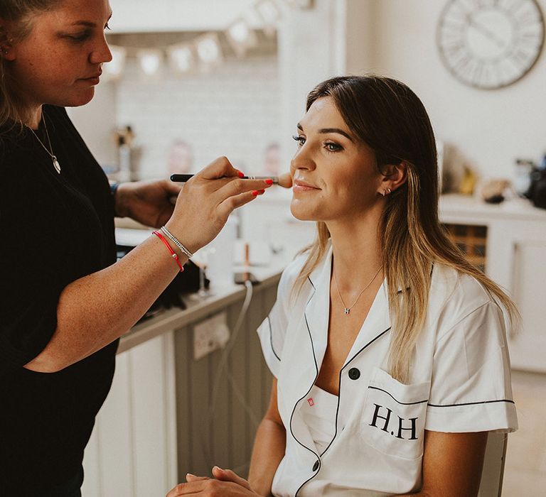 The bride in white satin monogrammed bridal pyjamas gets her makeup done for the wedding day 