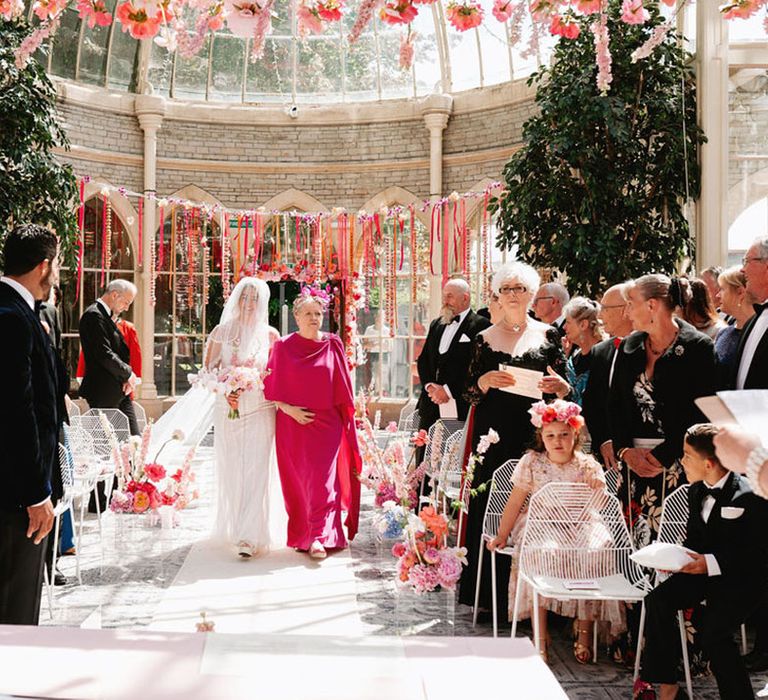 Mother of the bride in bright pink wedding outfit walking the bride down the aisle 