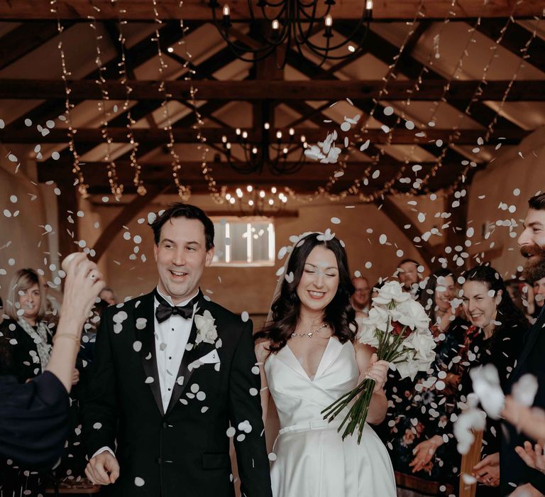 Bride holding white rose bridal bouquet and groom walk down the aisle with a confetti exit using monochrome confetti