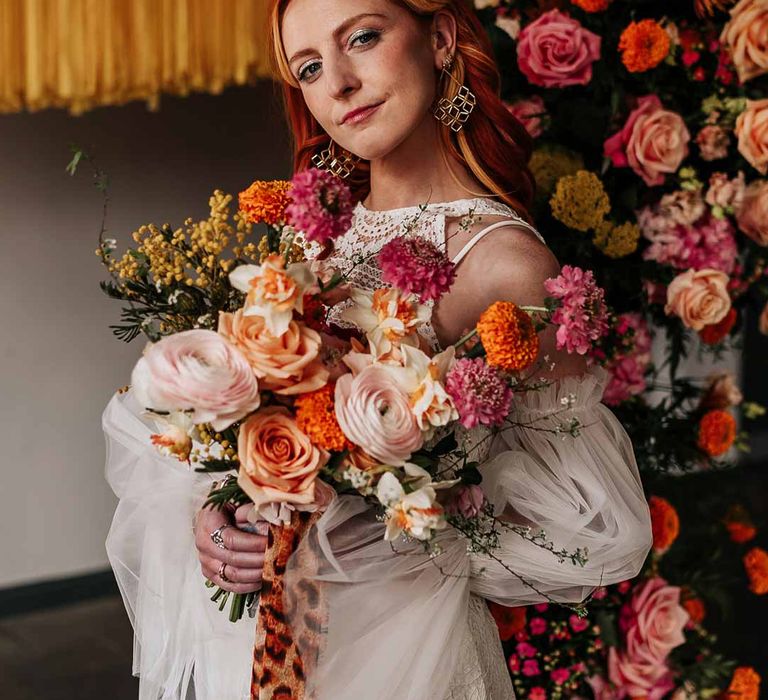 Bride in 70s-inspired boatneck short lace wedding dress with detachable tulle puff sleeves and platform gold wedding heels holding large colourful mixed flower bridal bouquet tied with leopard print ribbon 