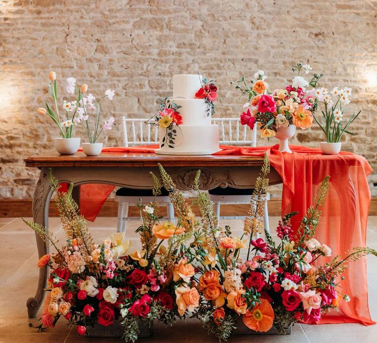 Merriscourt wedding venue reception room with wooden table with orange table runner and mixed seasonal flower arrangements and misshapen three tiered wedding cake with white icing and seasonal flower and foliage cake toppers 