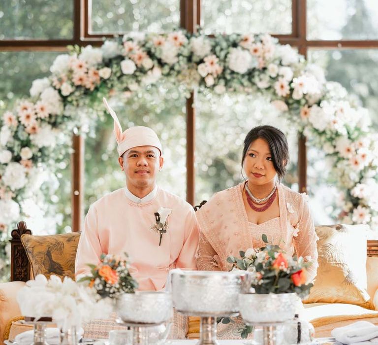 Bride in floral 3D applique pink and rose gold htamane sitting with groom in baby pink htaingmathein at Burmese wedding underneath floral wedding bouquet 