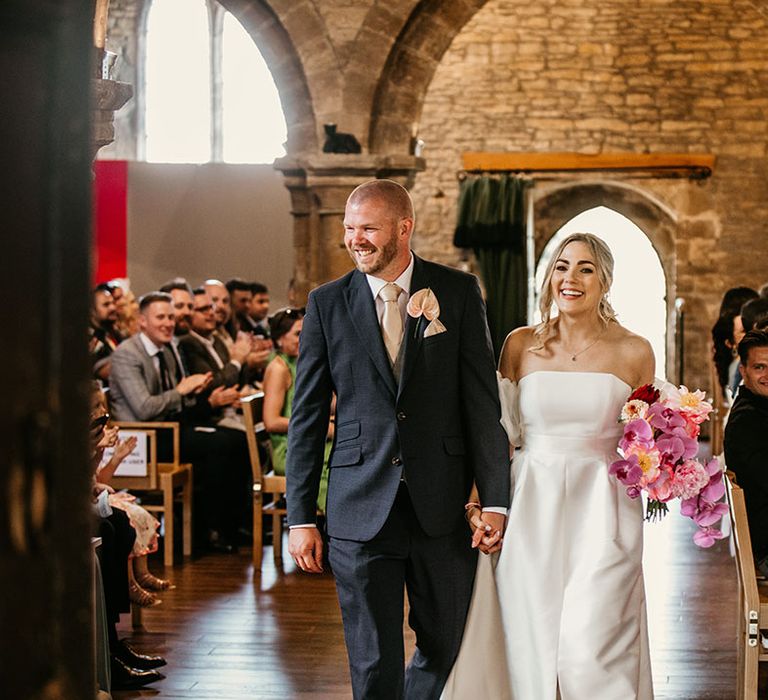 Bride in off the shoulder Jesus Peiro wedding dress with off the shoulder detail with the groom in a navy suit exiting from their church ceremony 