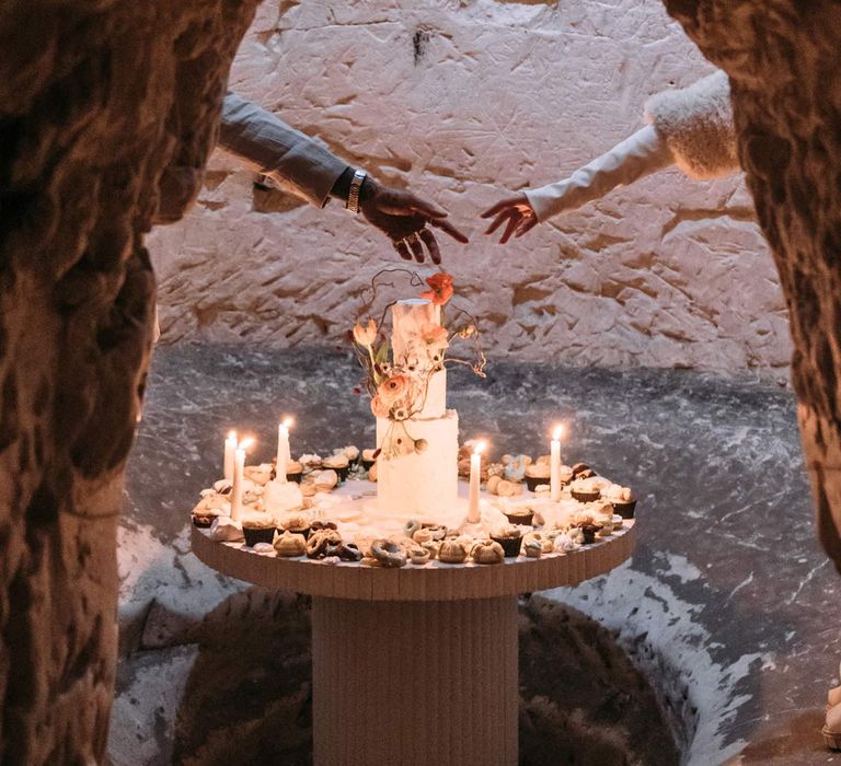 Bride and groom reaching their hands out to each other in front of three tiered iced wedding cake on wedding grazing table surrounded by tapered candles in Margate Caves