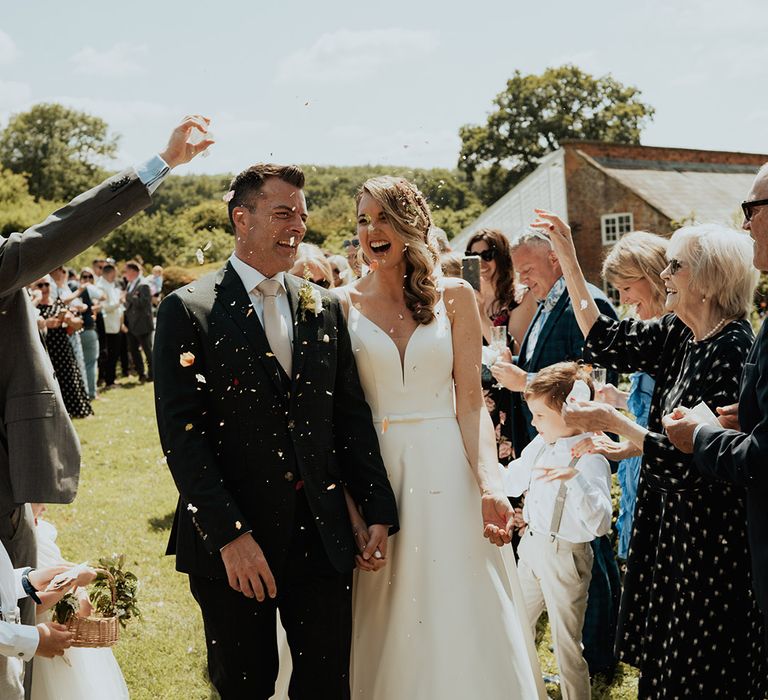 Bride in traditional white wedding dress with groom in suit with pale pink tie walking out of the ceremony to confetti 