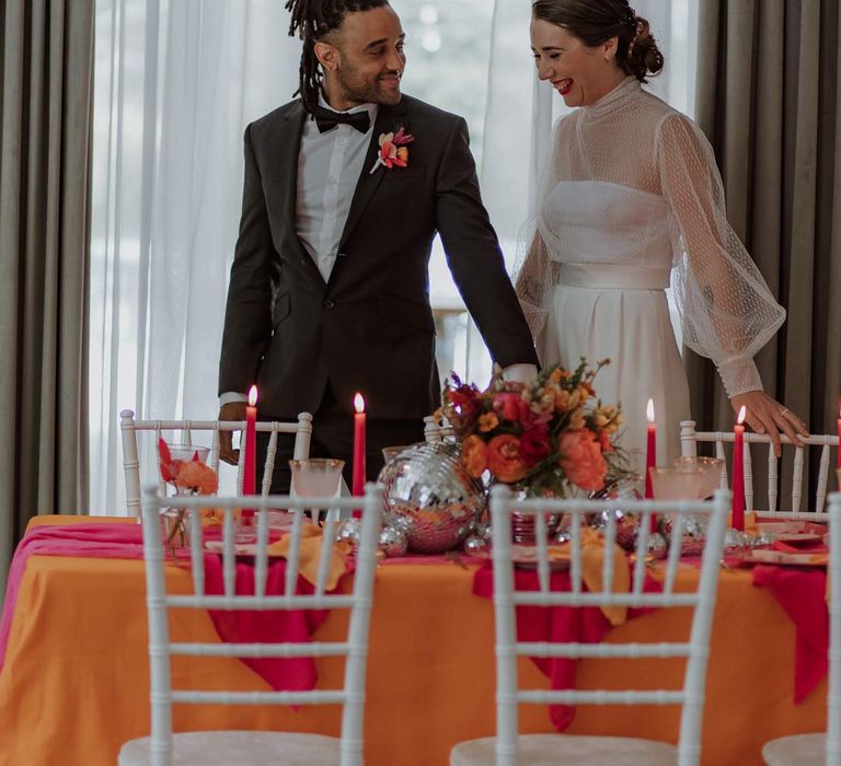 Groom in classic black tuxedo with bowtie and pink and orange wedding boutonniere with bride in mesh long sleeve high neck polka dot wedding dress stood by pink and orange wedding tablescape with disco ball decor and bright floral centrepieces 