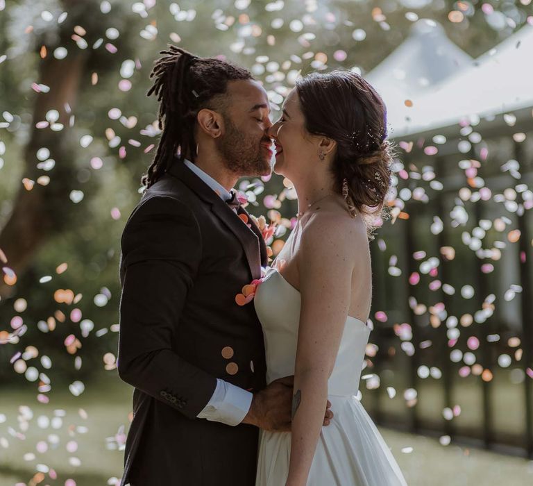 Bride in strapless sweetheart neckline wedding dress holding pink and orange wedding bouquet with orange ribbon kissing groom in classic black tuxedo with bowtie and pink and orange wedding boutonniere with colourful wedding confetti falling around them