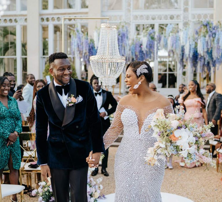 Bride in illusion v-neck sheer off shoulder long sleeve pearl embellished wedding gown and groom in velvet midnight blue double breasted groom’s blazer, velvet bowtie and peach garden rose boutonniere walking down the aisle at Syon Park vow renewal 