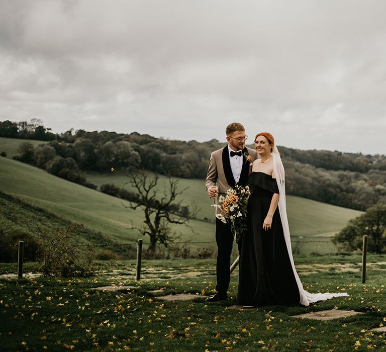 Bride in black satin off the shoulder wedding dress with groom in field of Surrey hills 