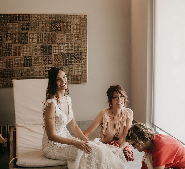 bride in her wedding morning in an appliqué wedding dress with her bridal party helping her put on her wedding shoes 