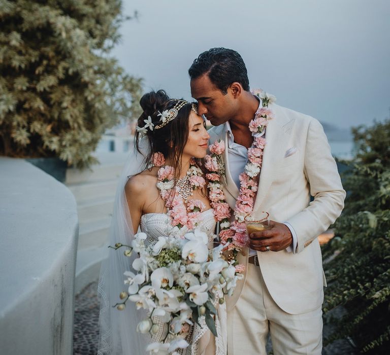 Boho bride in white lace wedding dress with flower hair accessories and traditional Matha Patti holding a white orchid wedding bouquet with groom in groom suit and both wearing traditional pink Varmalas