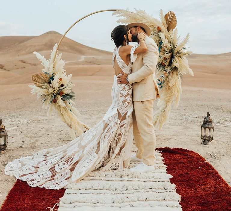 Bride in boho lace wedding dress kisses the groom in front of the boho moongate decoration 