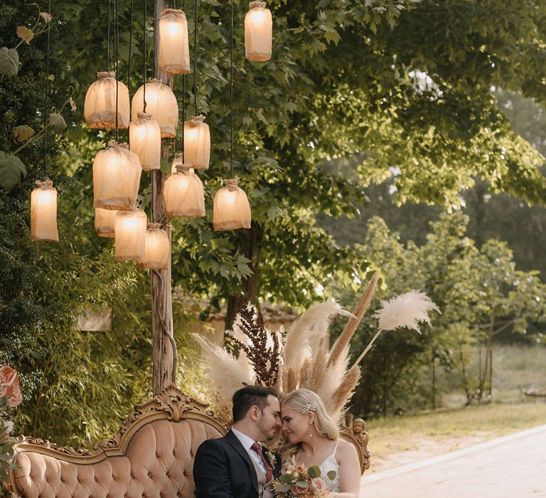 Bride and groom sitting on sofa seating area with rustic rattan lighting decorations hanging above 