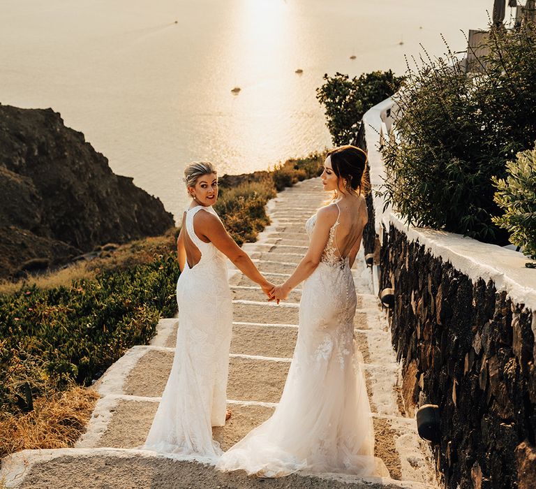 Two brides wearing boho wedding dress with lace detailing and open backs as they walk hand in hand at golden hour 