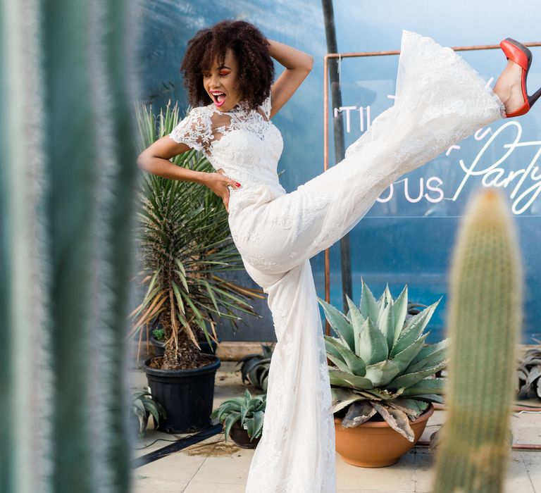 Bride in white Lillian West Jumpsuit and red heels kicking leg in front of neon sign