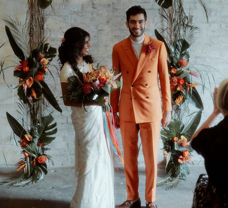 Bride in a white sari and groom in an orange suit standing at the altar decorated in tropical flowers