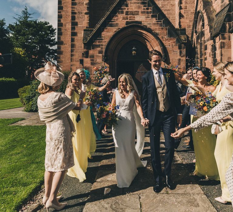 Bride in Justin Alexander wedding dress holding colourful bouquet holds hand with groom whilst wedding guests throw confetti outside of church