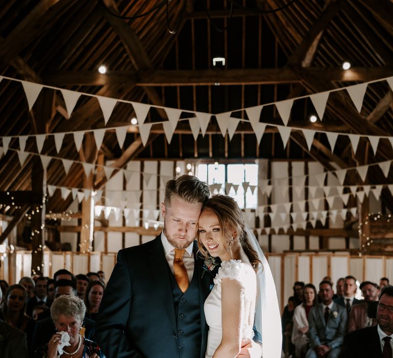 Bride in a fitted wedding dress with cathedral length veil embracing her husband in a navy suit at the altar 