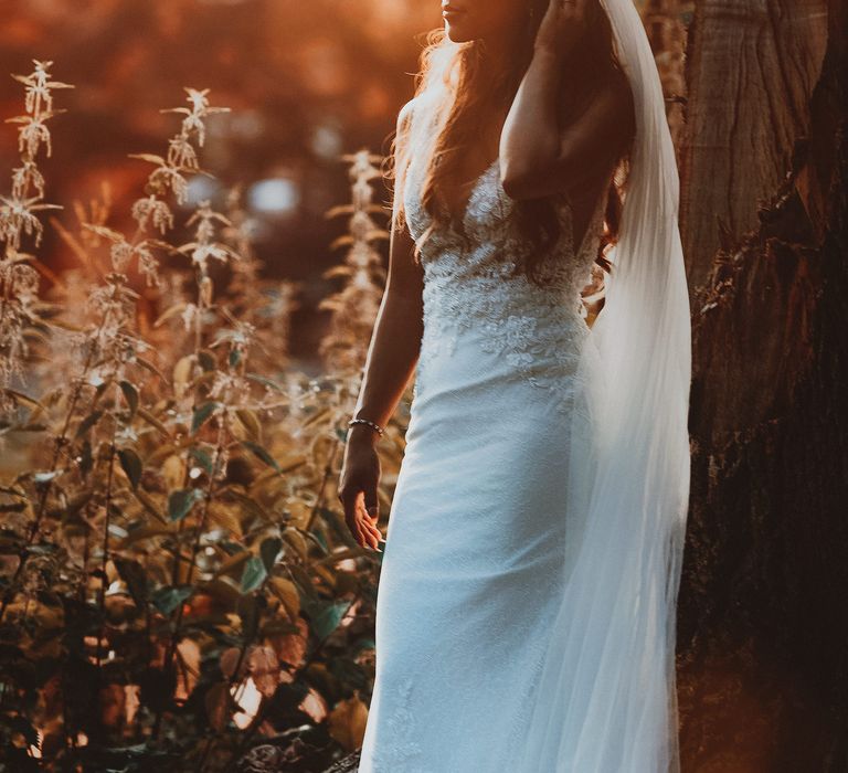 Bride stands outdoors in bridal gown with light behind her