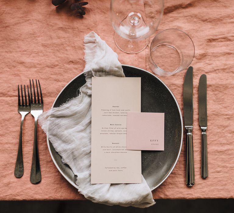 Place setting with dusky pink tablecloth, black charger plate and beige menu card 