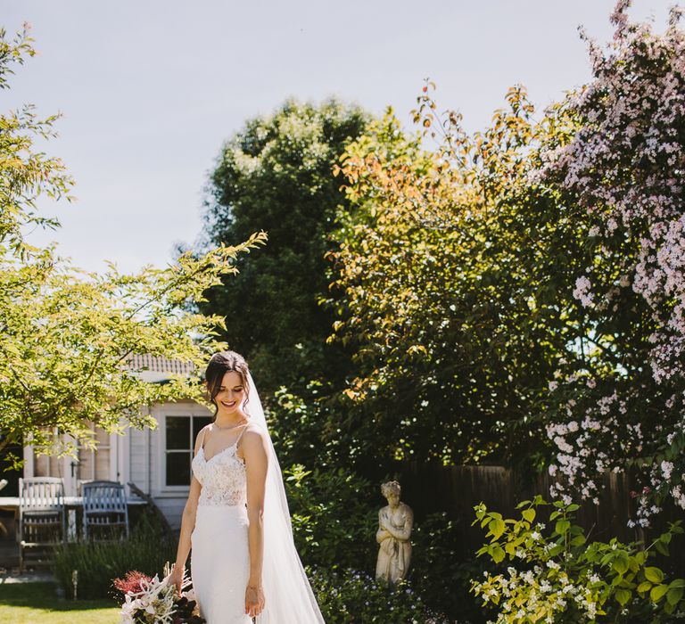 Dark haired bride stands in garden wear Enzoani wedding gown and carrying floral bouquet