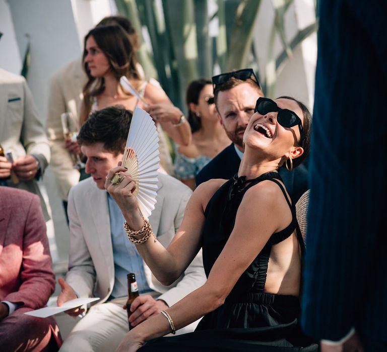 Wedding guest laughing and fanning herself, wearing black sunglasses and a black sun dress