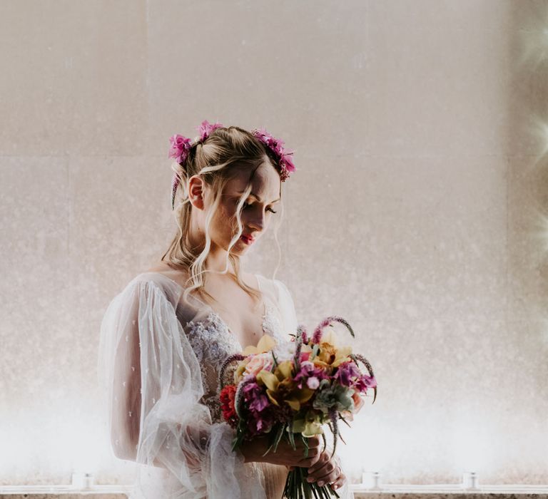 Bride with curled hair, pink floral hair accessories and long tulle sleeved boho wedding dress holds multicoloured wedding bouquet whilst standing in front of fairy lights at enchanted forest wedding in Italy