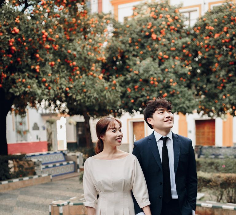 Asian couple hold hands in Seville plaza in front of fountain and orange trees