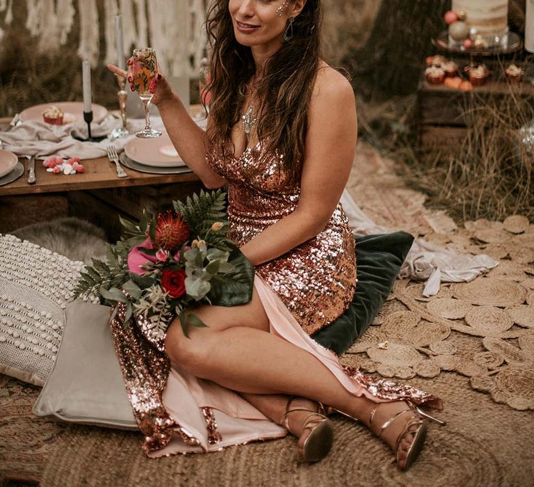 Bride with long curled hair in rose gold sequin dress with slit and face glitter holds multicoloured bouquet and sparkling wine whilst sat on cushion at festival themed wedding