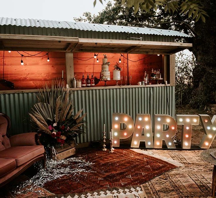 Outdoor bar with rugs, velvet sofa and lightbulb PARTY sign at festival themed wedding