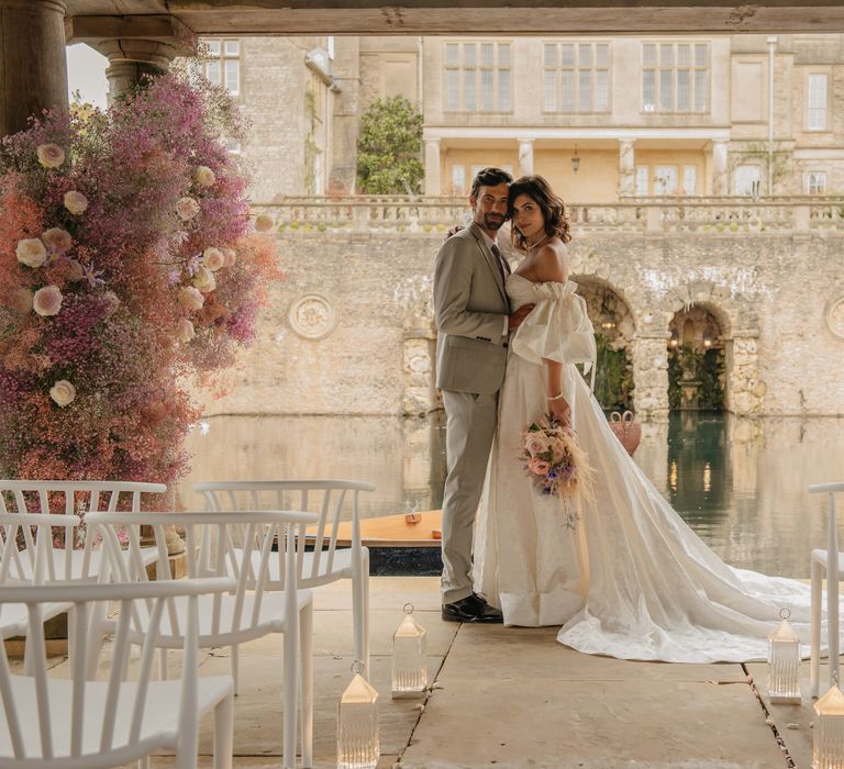 Bride & groom stand at the front of aisle whilst Italian style lake and archways can be seen in the background