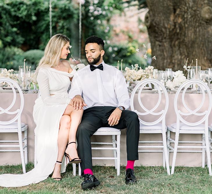 Sophisticated bride and groom sitting at their outdoor wedding reception on white chairs 