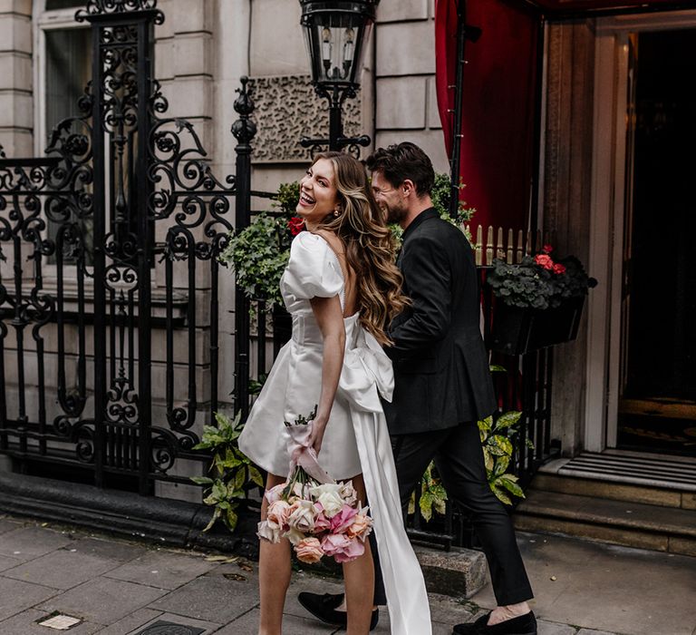 Portrait of a bride in a short wedding dress with long trailing bow back detail walking through Mayfair with her groom 