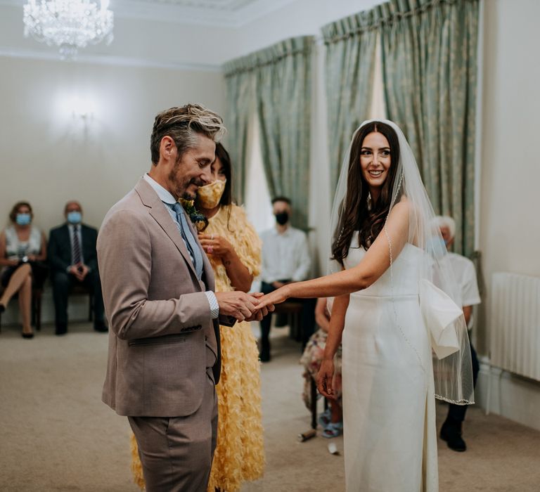 Bride in white strapless Rebecca Vallance Dress, and veil smiles whilst groom in brown Moss Bros suit and blue tie puts ring on her finger as guest in yellow dress looks on at Harrogate wedding 