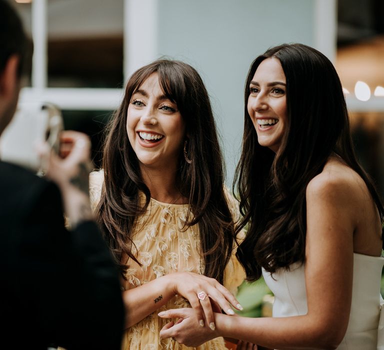 Bride in white strapless Rebecca Vallance dress stands having polaroid taken with wedding guest in midi yellow short sleeved dress with feather detailing in courtyard at Hotel du Vin wedding reception in Harrogate