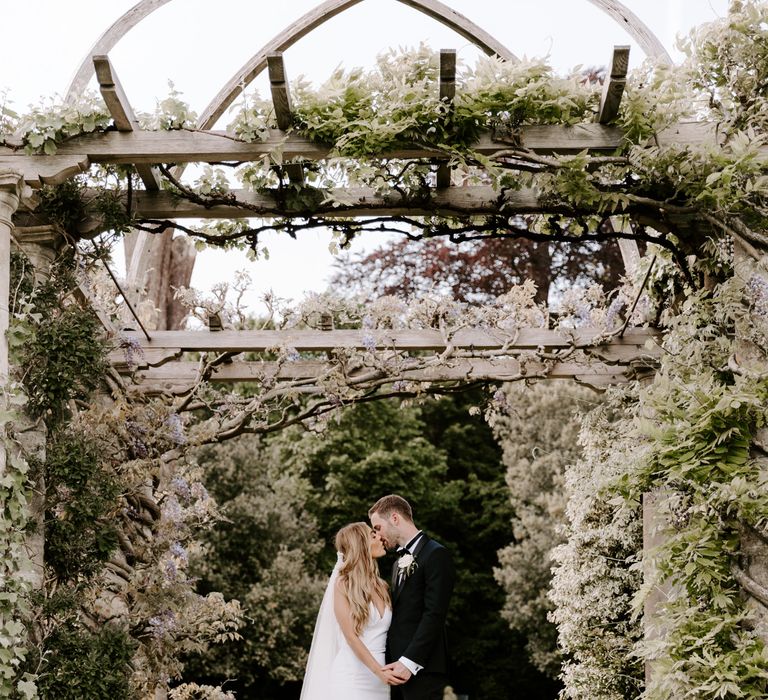 Bride & groom stand beneath pergola outdoors on their wedding day