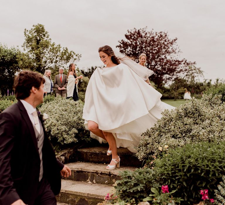 Bride in white Elbeth Gillis wedding dress and pale grey block heels walks down garden steps whilst holding up dress at home farm wedding