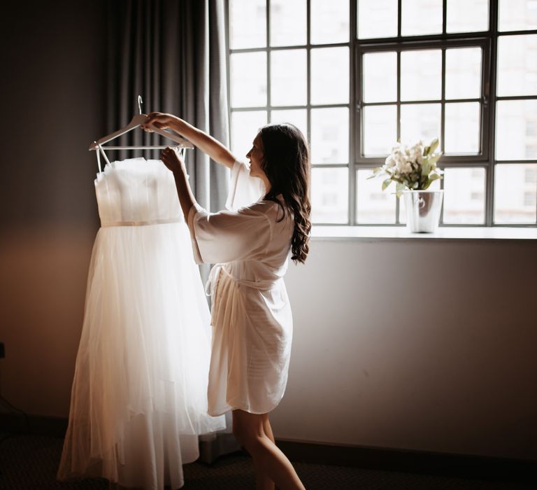 Bride looks at wedding gown in front of window on the morning of her wedding day