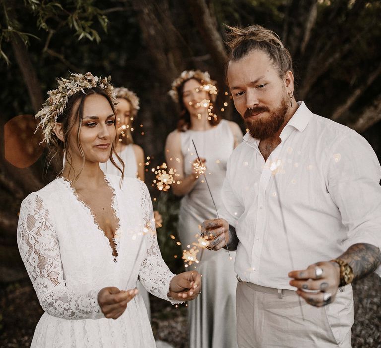 Boho bride and groom sparkler moment with bride in a lace wedding dress and dried flower crown and groom in a white skirt and beige trousers 