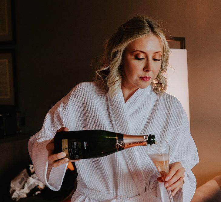 Bride in a white waffle getting ready robe pouring champagne on the wedding morning 