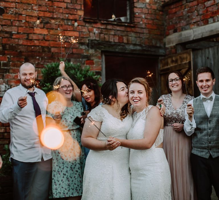 Bride kisses her bride on the cheek as they are surrounded by wedding guests holding sparklers outdoors