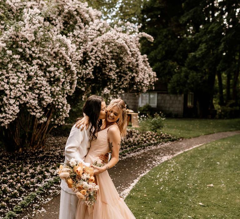 Bride in a white suit kissing her brides cheek in a blush pink wedding dress as she holds a yellow flower bouquet 