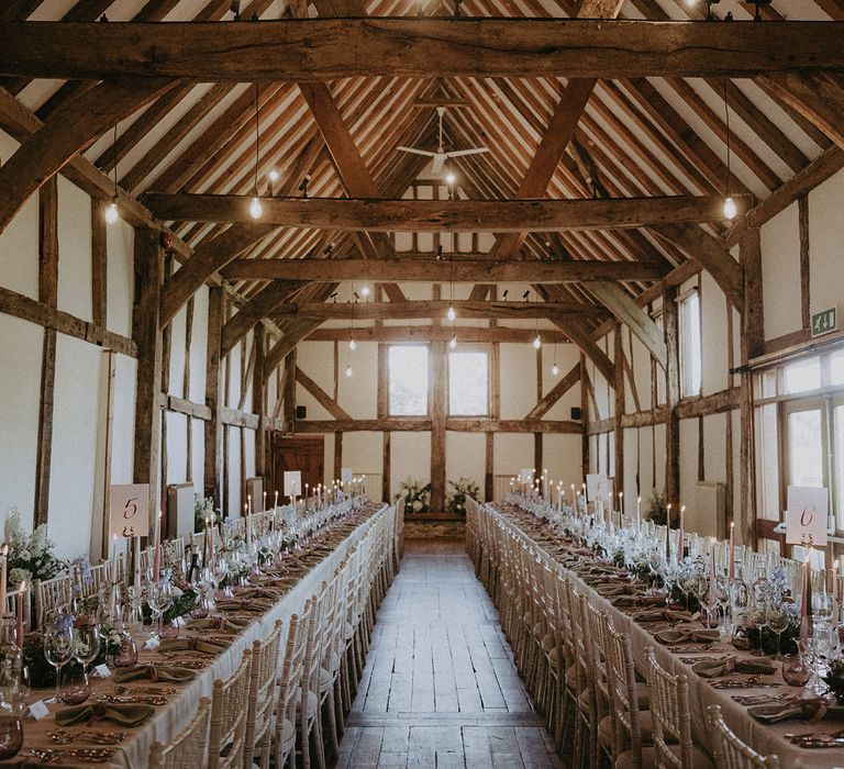 Two long wedding breakfast tables with white bamboo style chairs, lit candles and glassware with pink and green details, wooden beams and wooden floor inside Loseley Park