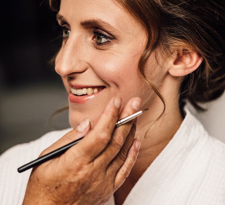 Smiling bride with wedding updo in dressing gown gets her make up done before summer wedding in Dorset 